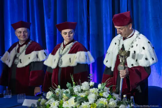 Trzej mężczyźni w togach akademickich z łańcuchami, przy stole z kwiatami.