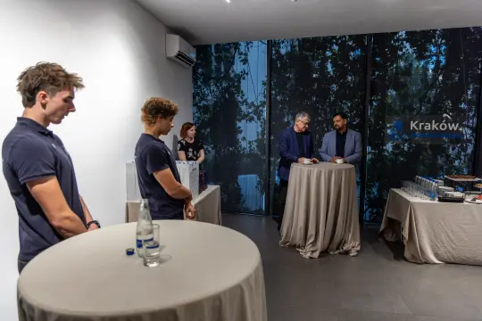 Spotkanie w eleganckim wnętrzu z dwoma mężczyznami przy stole i grupą osób w tle.