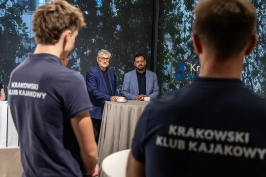 Dwóch mężczyzn przy stole, w tle osoby w koszulkach z napisem Krakowski Klub Kajakowy.