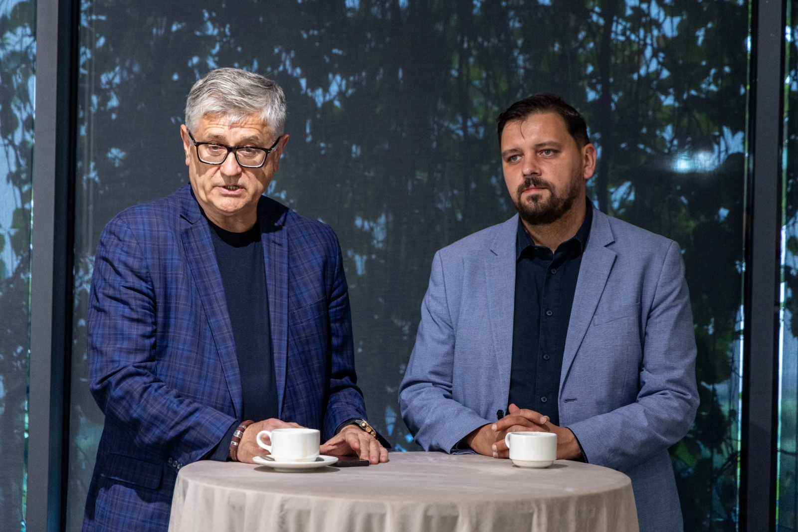 Dwóch mężczyzn przy okrągłym stole, jeden z filiżanką kawy, rozmawia w jasnym wnętrzu.  Autor: Jan Graczyński