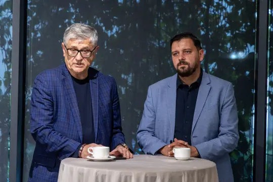 Dwóch mężczyzn przy okrągłym stole, jeden z filiżanką kawy, rozmawia w jasnym wnętrzu.