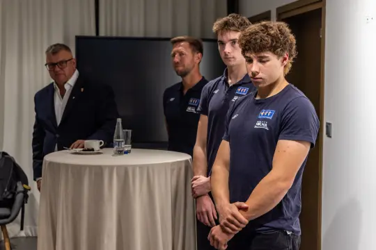 Czterech mężczyzn w strojach sportowych, dwóch z nich stoi z poważnymi minami, jeden z boku obserwuje.