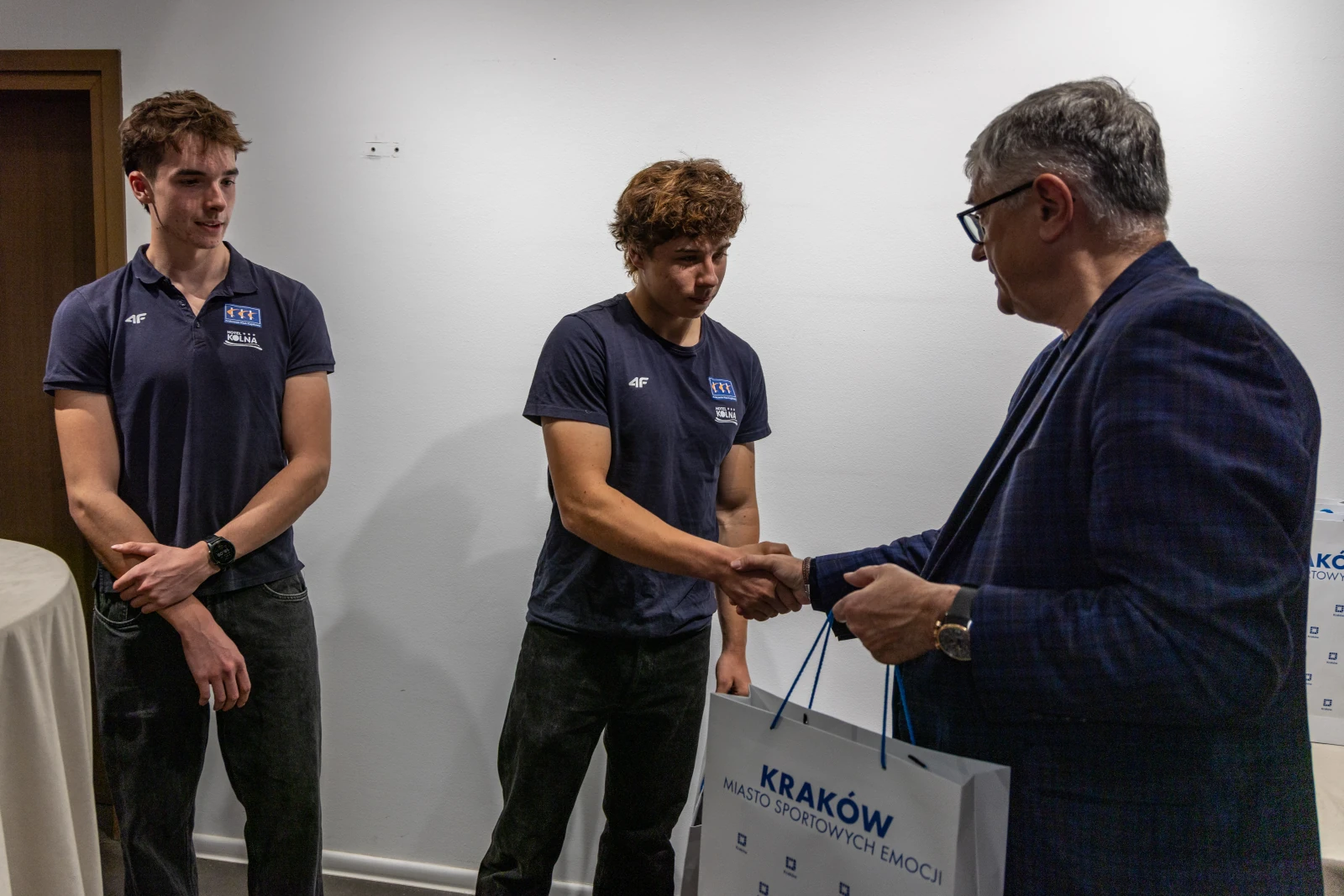Dwóch młodych sportowców w rozmowie z mężczyzną w okularach, wręczającym torbę.  Autor: Jan Graczyński