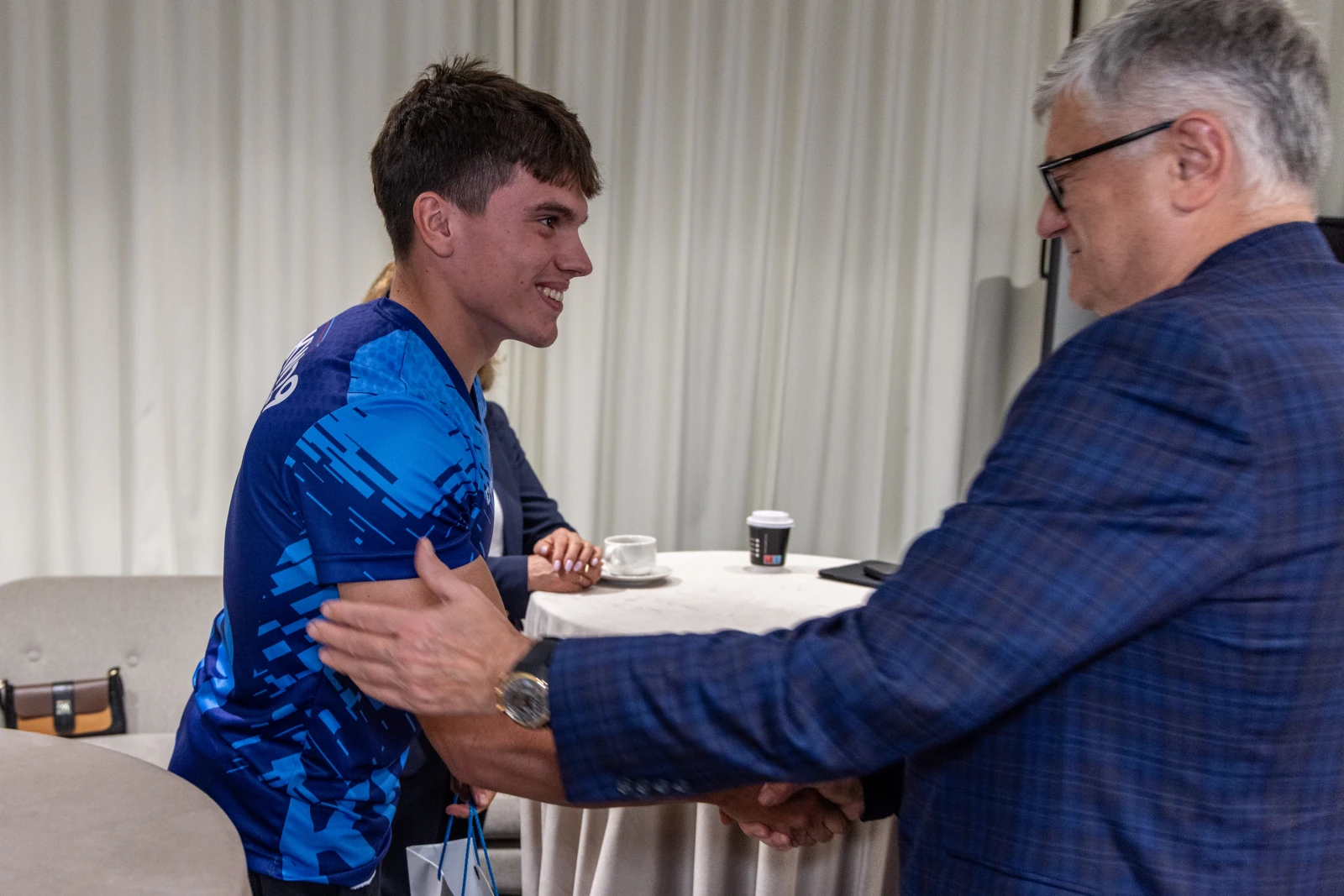 Młody sportowiec uścisnął dłoń starszemu mężczyźnie w formalnym otoczeniu.  Autor: Jan Graczyński