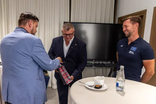 Trzech mężczyzn w formalnych strojach, jeden wręcza prezent, na stole kawa i ciasto.