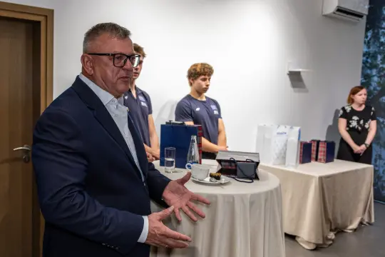 Mężczyzna w garniturze przemawia, w tle młodzi ludzie i stoły z prezentami.