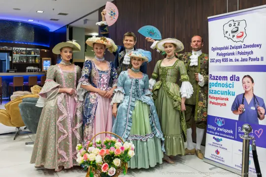 Grupa osób w strojach historycznych, w tle baner promujący pielęgniarki i położne.