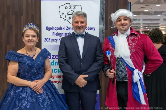 Trzy osoby w eleganckich strojach, w tym kobieta w niebieskiej sukni i mężczyzna w czerwonym płaszczu.