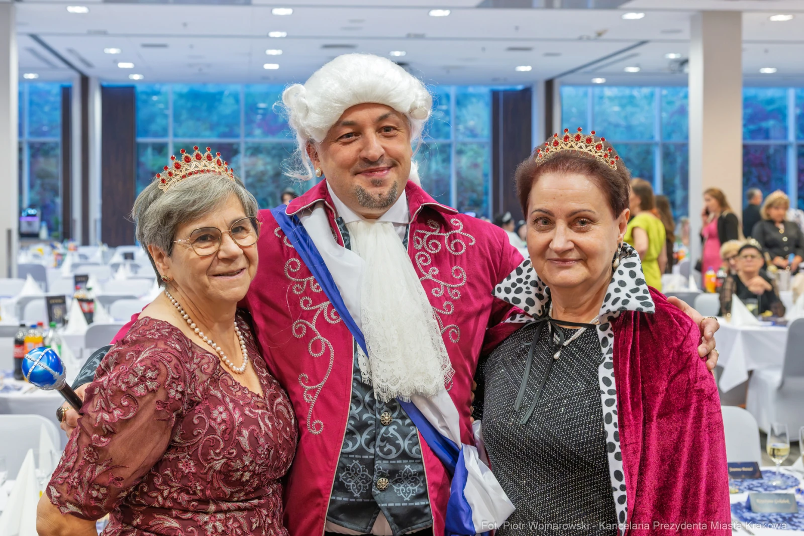 Trzy osoby w eleganckich strojach, jedna w koronie, w sali bankietowej.  Autor: Piotr Wojnarowski
