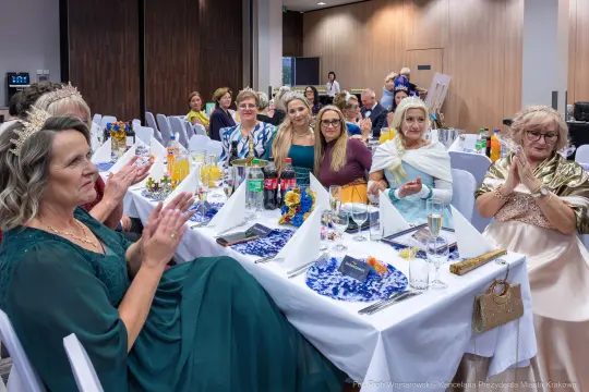 Grupa kobiet w eleganckich strojach bawi się na przyjęciu, siedząc przy stołach z jedzeniem i napojami.