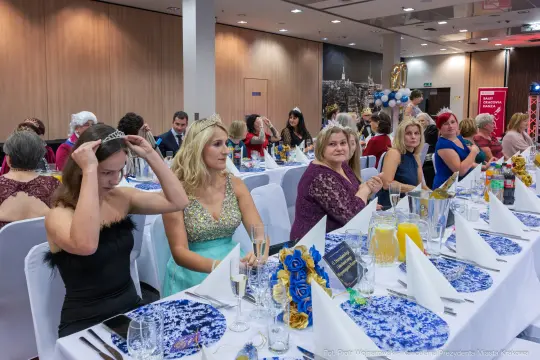 Kobiety w eleganckich strojach siedzą przy stole na uroczystości, w tle dekoracje i goście.