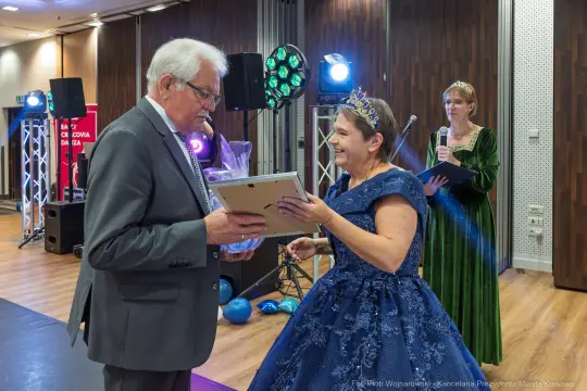 Mężczyzna wręcza nagrodę kobiecie w niebieskiej sukni, w tle dekoracje i oświetlenie.