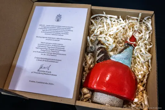 Ceramiczna figurka w kształcie koguta w czerwonym kapeluszu, obok list w eleganckim pudełku.