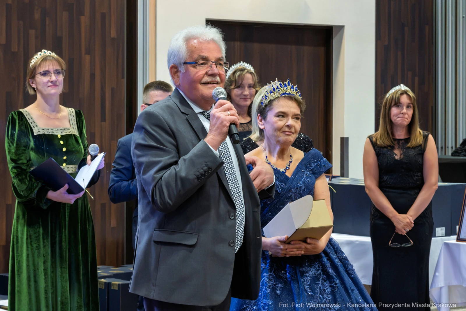 Mężczyzna przemawia, obok niego kobieta w niebieskiej sukni z koroną, w tle inni goście w eleganckich strojach.  Autor: Piotr Wojnarowski