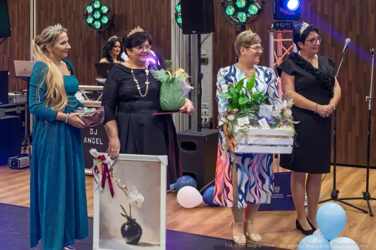 Cztery kobiety w eleganckich strojach z nagrodami i kwiatami na scenie podczas uroczystości.