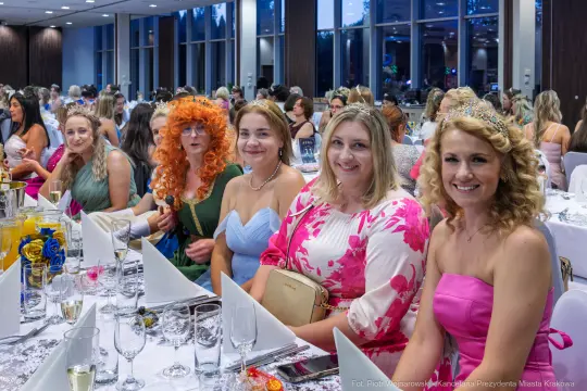 Grupa kobiet w eleganckich strojach, uśmiechających się przy stole na przyjęciu.