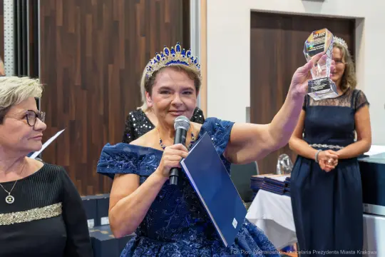 Kobieta w niebieskiej sukni z koroną trzyma statuetkę i przemawia do zgromadzonych gości.