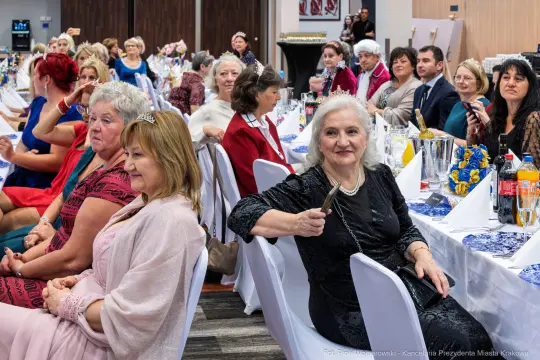 Grupa kobiet i mężczyzn w eleganckich strojach na uroczystości, przy stołach z jedzeniem i napojami.