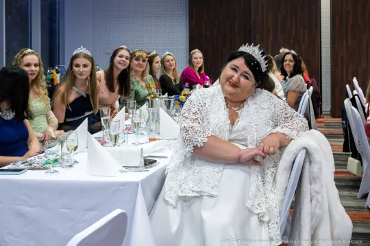 Kobieta w białej sukni i koronie siedzi przy stole, wokół niej goście w eleganckich strojach.