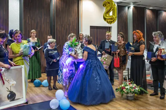 Uroczystość z wręczeniem nagród, kobiety w eleganckich strojach, balony i kwiaty.