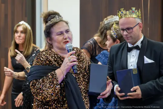 Kobieta w eleganckiej sukni z mikrofonem, obok mężczyzna w garniturze i koronie, inni goście w tle.