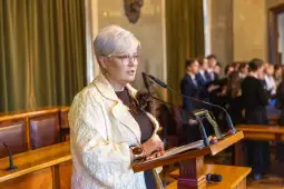 Kobieta w okularach przemawia z mównicy, obok niej stoi kot. W tle grupa młodych ludzi.