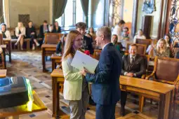 Uroczystość wręczenia nagrody, uśmiechnięta kobieta z różą i mężczyzna z dokumentem.
