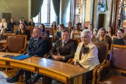 Grupa ludzi siedzących w sali, w tym trzech mężczyzn i kobieta, w formalnych strojach.
