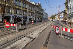 Zamknięta ulica z barierkami i torami tramwajowymi, w tle budynki i pojazdy.