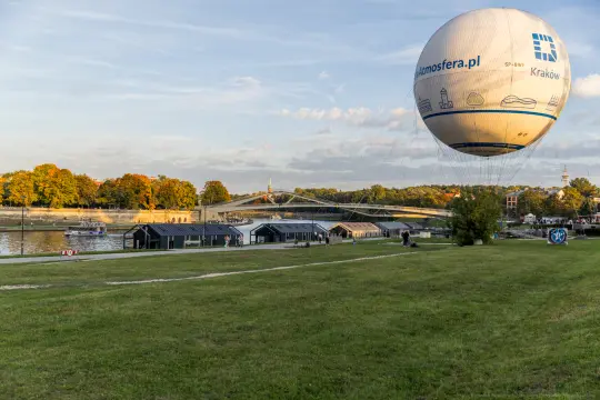 Balon na ogrzane powietrze nad rzeką, w tle most i zielona przestrzeń.