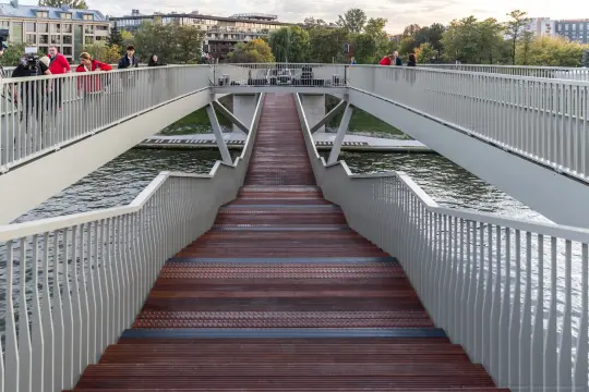 Nowoczesny most z drewnianymi schodami nad wodą, otoczony zielenią i ludźmi.