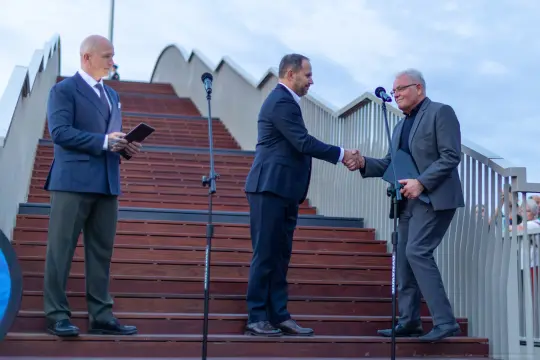 Trzech mężczyzn na schodach, dwóch z nich podaje sobie ręce, trzeci obserwuje.