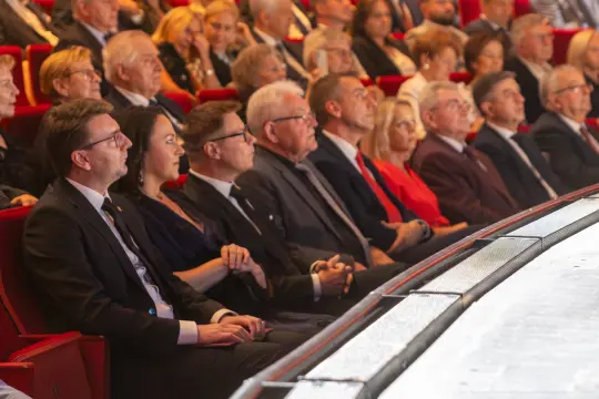 Grupa ludzi w eleganckich strojach siedzących na widowni w teatrze.
