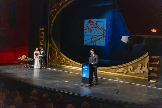 Mężczyzna przemawia z podium na tle dekoracji teatralnej, obok dwie osoby w formalnych strojach.