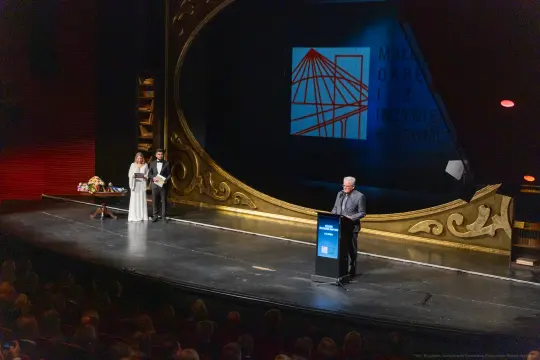 Mężczyzna przemawia na scenie, obok dwie osoby, w tle logo wydarzenia.