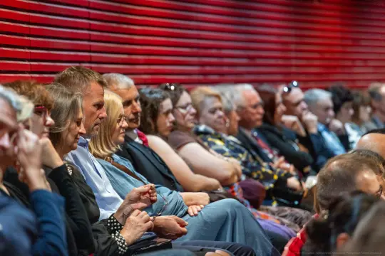Grupa ludzi siedzących w rzędach na wydarzeniu, skupionych na prezentacji.