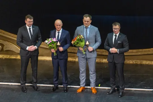 Czterech mężczyzn w garniturach trzymających kwiaty na scenie teatralnej.
