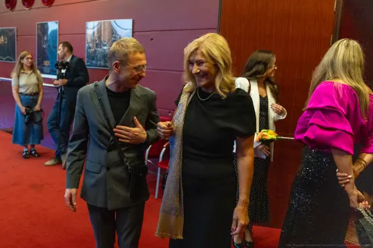 Uśmiechnięta para rozmawia w eleganckim wnętrzu, w tle inni goście.