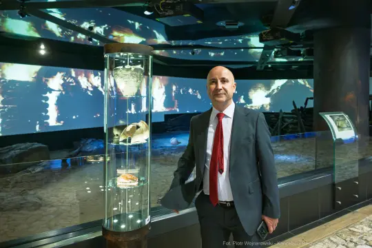 Mężczyzna w garniturze stoi obok szklanej gabloty z eksponatami w muzeum. W tle projekcja multimedialna.