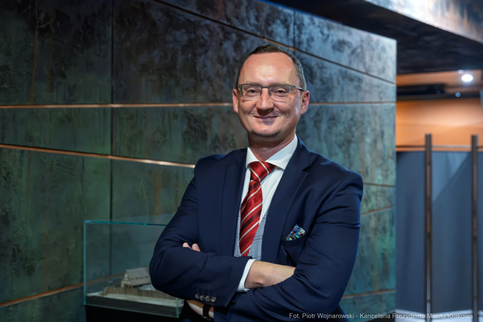 Mężczyzna w garniturze z uśmiechem, stojący z założonymi rękami w nowoczesnym wnętrzu.  Autor: Piotr Wojnarowski