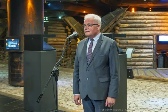 Mężczyzna w garniturze stoi przy mikrofonie w nowoczesnym wnętrzu.
