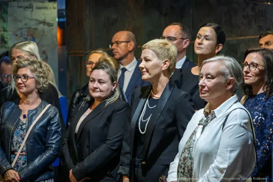 Grupa osób w formalnych strojach, zróżnicowane wyrazy twarzy, w tle industrialne wnętrze.