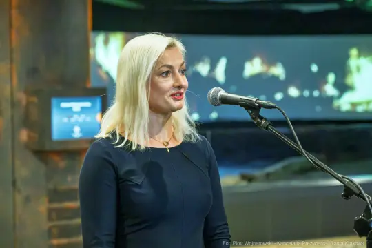 Kobieta z blond włosami przemawia do mikrofonu w tle z projekcją.