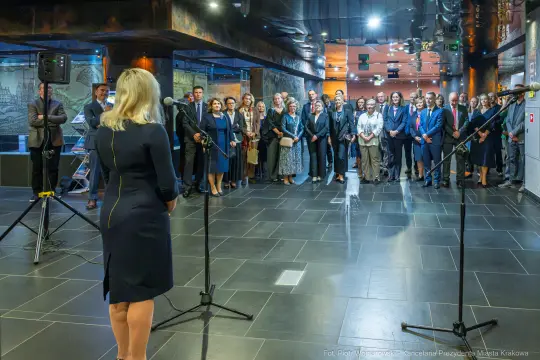 Uczestnicy wydarzenia stoją w sali, słuchając przemówienia.