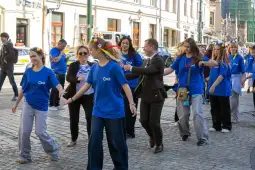 Grupa osób w niebieskich koszulkach tańcząca na ulicy, część z kwiatowymi wiankami na głowach.