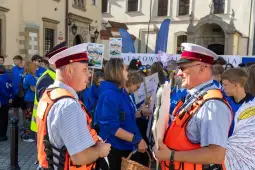 Dwaj mężczyźni w mundurach rozmawiają, w tle grupa młodzieży z plakatami.