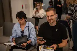 Kobieta z gazetą i mężczyzna z telefonem w sali pełnej ludzi.