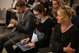 Grupa ludzi siedzących na spotkaniu, jedna osoba pracuje na laptopie.