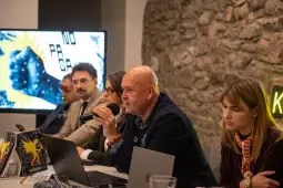 Mężczyzna z mikrofonem prowadzi dyskusję przy stole, w tle prezentacja na ekranie.