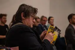 Mężczyzna z okularami trzyma w rękach gazetę, siedząc wśród innych osób.